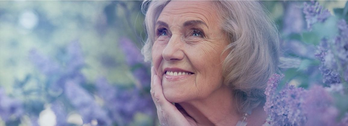 Ältere Person mit hellblonden Haaren, die eine Hand an das Kinn legt, vor unscharfem Hintergrund mit lila Blüten.